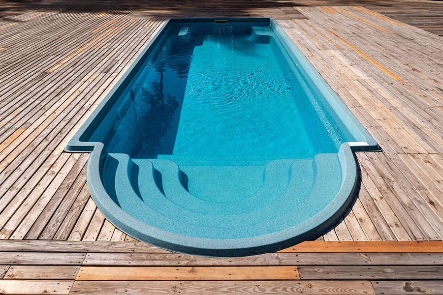 New modern fiberglass plastic swimming pool entrance step with clean fresh refreshing blue water on bright hot summer day at yard or resort hotel spa area. Wooden flooring deck of teak or larch board.